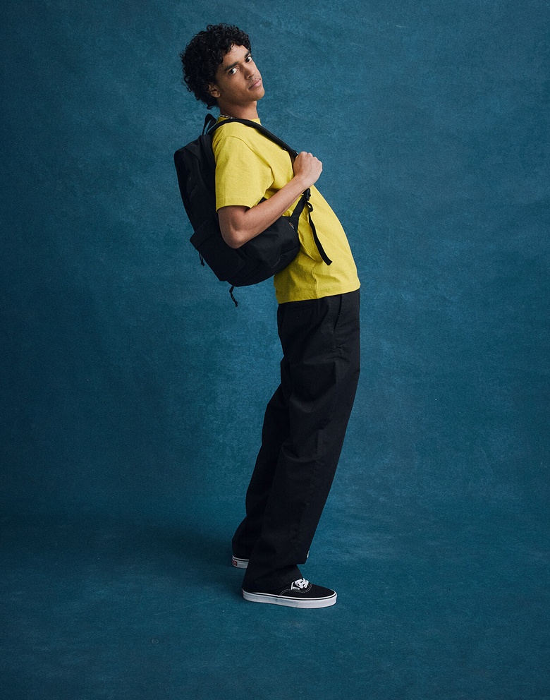 A young man posing against a picture day background in black and white Authentic, a black Old Skool Trek Backpack, and a yellow Stacked Hi T-Shirt.