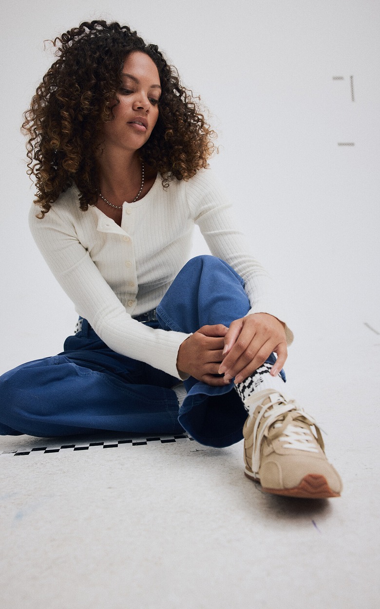 A young woman sitting on the floor of a photography studio wearing incense brown Premium Super Lowpro Trainers and Check-5 Low Rise Baggy Pants.