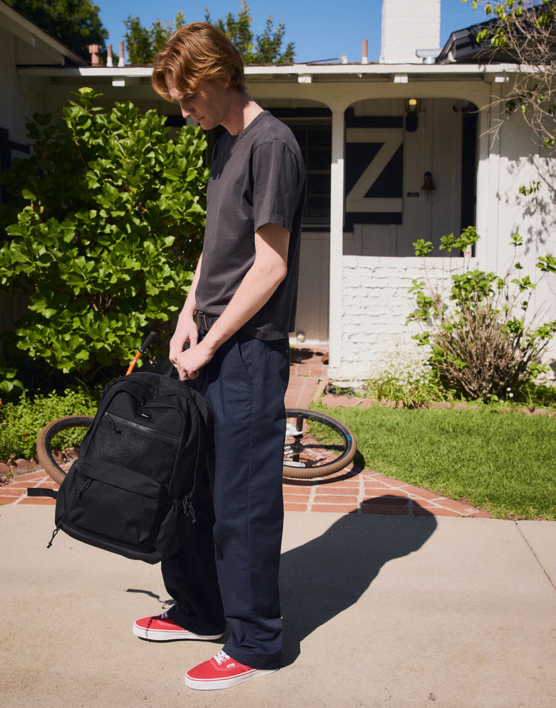 A young man outside of a house wearing red Authentic, a black Old Skool Trek Backpack, and Parisian night blue Authentic Chino Loose Pants.