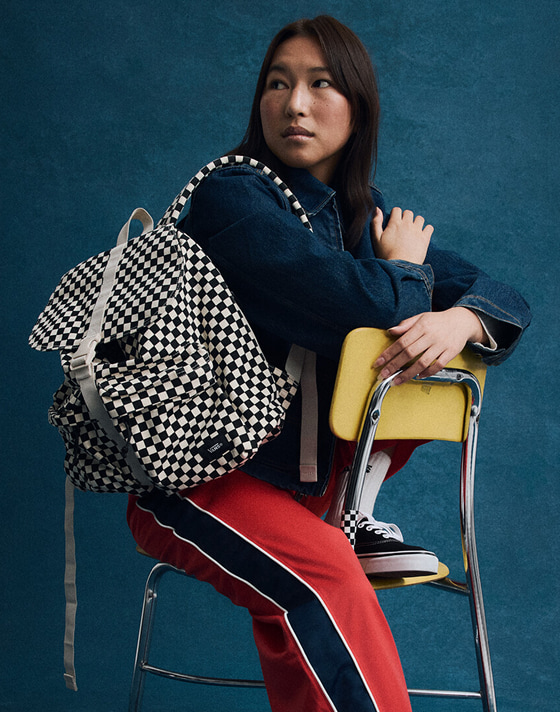 A young woman sits on a yellow classroom chair wearing a black and white Checkerboard Scatter Backpack.