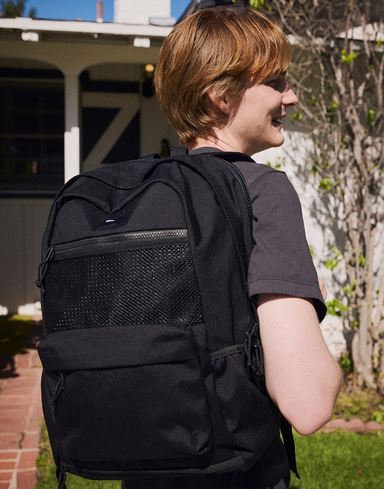 A young man sits on a bike outside of his house wearing a black Divert Backpack.