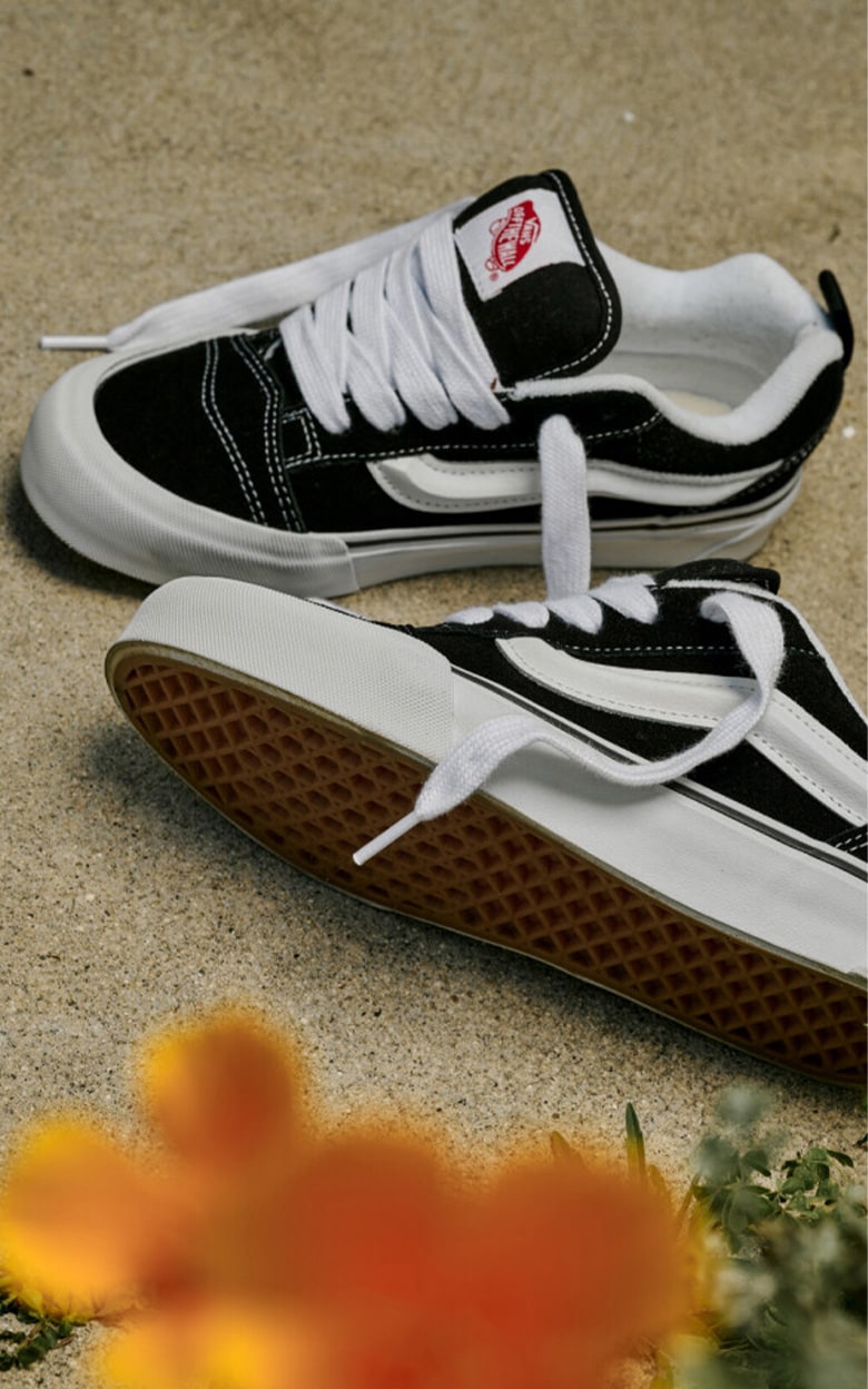 A pair of black Knu Skool shoes with laces untied rest on a concrete background with yellow flowers growing below.