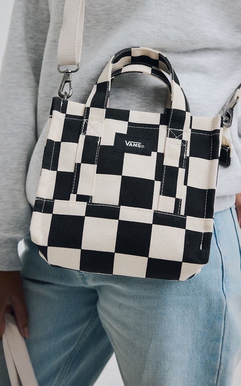 Un sac fourre-tout Checkerboard Lil Pergs noir et blanc est appuyé contre la hanche d’une personne portant un chandail gris et un jean bleu.
