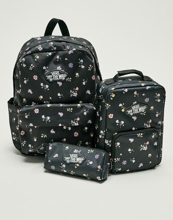 A black floral Old Skool Grom Backpack, Old Skool Lunch Bag, and Old Skool Pencil Pouch on the floor of a photography studio.