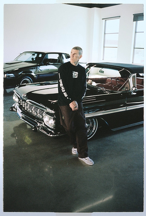 Image of Travis Barker in a car garage leaning against a car and wearing the Vans x Travis Barker Old Skool shoes.