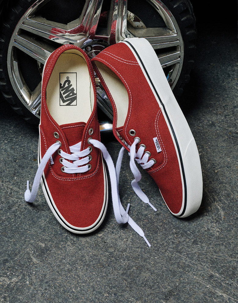 A pair of red Authentic shoes lean up against the silver hubcap of a car with their laces undone down to the concrete.