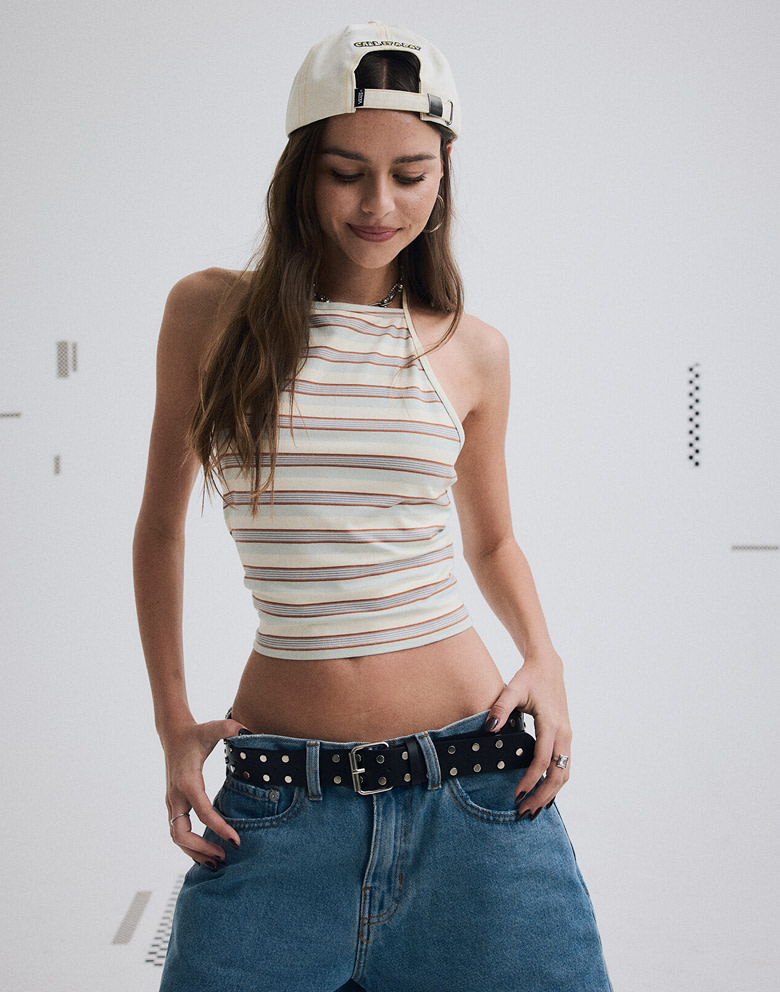 Waist up medium shot of a woman wearing a backwards hat, a crop top, a black belt, and baggy blue jeans.