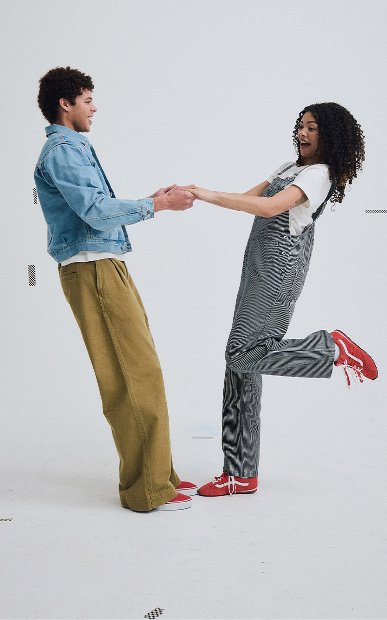 Medium shot of a man and a woman holding hands and leaning back in a grey photography studio dressed head to toe in Vans clothing and shoes.