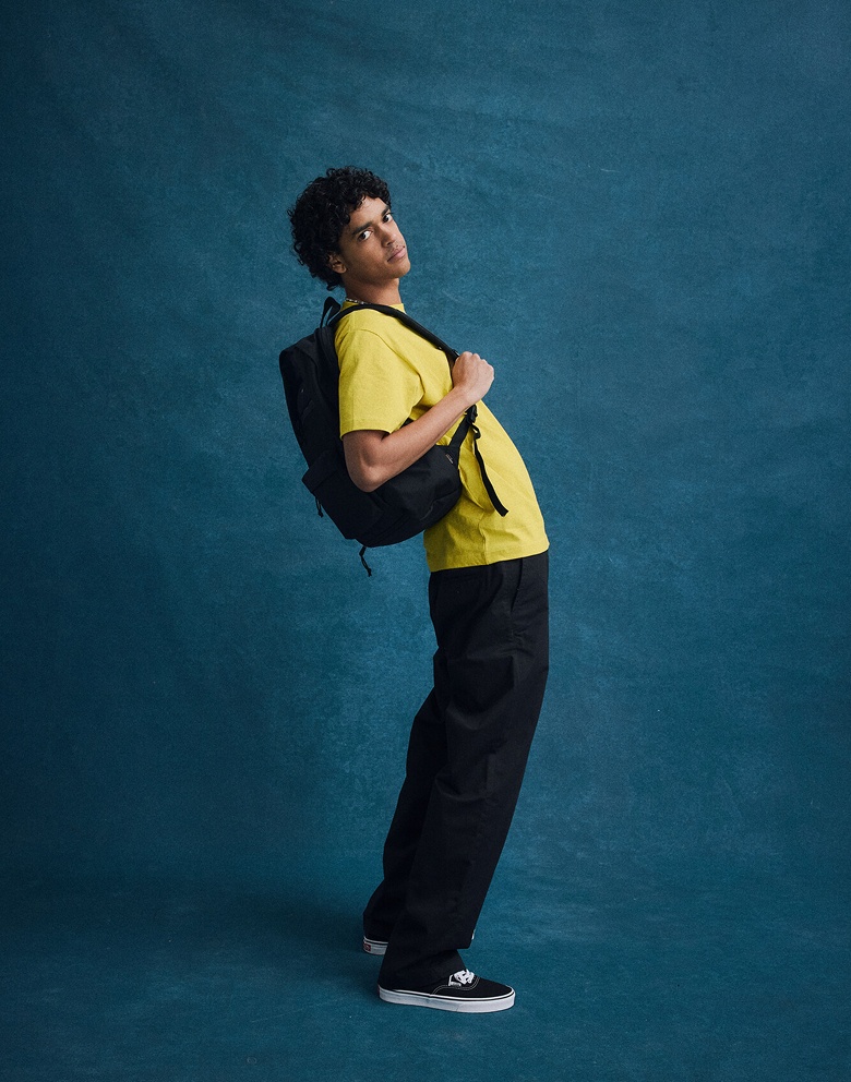 Wide shot of a man in a school photo setting with a blue backdrop. He’s wearing a yellow shirt, black pants, a black backpack and Authentic shoes.