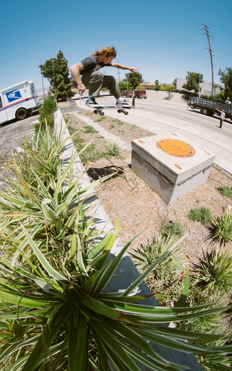 Panoramic shot of Vans Skateboarder T-FUNK. He’s mid-air over a wall of the Post Office parking lot and wearing the Checkerboard black/white Skate Era