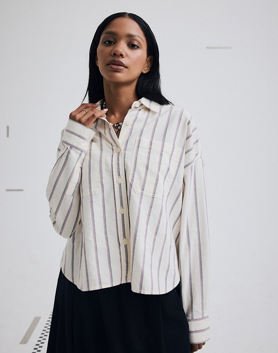 Medium shot of a woman waist up wearing the Long Sleeve Boxy Oxford Shirt in Egret White. She’s adjusting her collar in a grey photography studio.