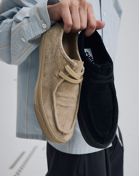Close up of a man’s hand holding one light brown, and one black Premium SF Moc Low Shoe in a grey photography studio.