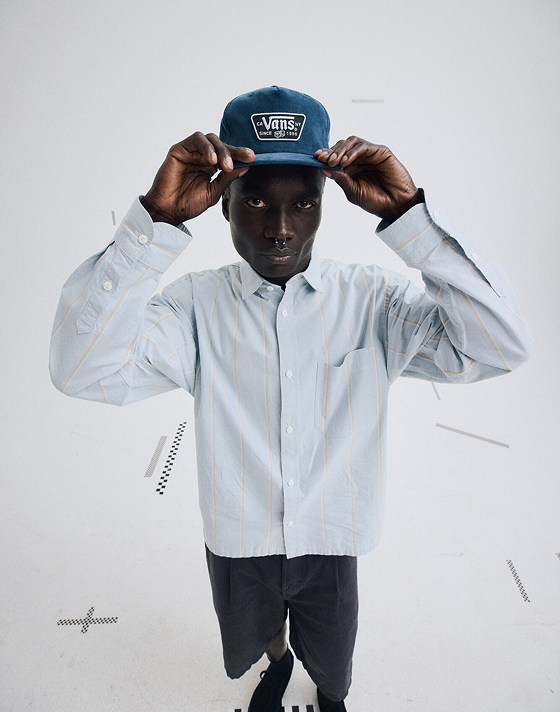 Wide shot in a grey photography studio of a man wearing a Blue Knu Patch Unstructured Hat and the Striped Oxford Long Sleeve Shirt in Soft Slate Blue.