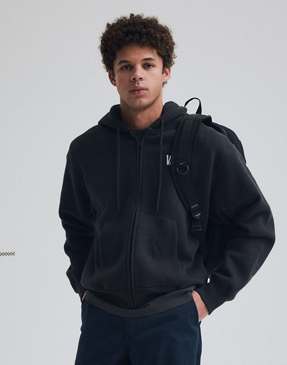 A young man poses in a photography studio in Vans clothes with a Vans backpack slung over his shoulder.