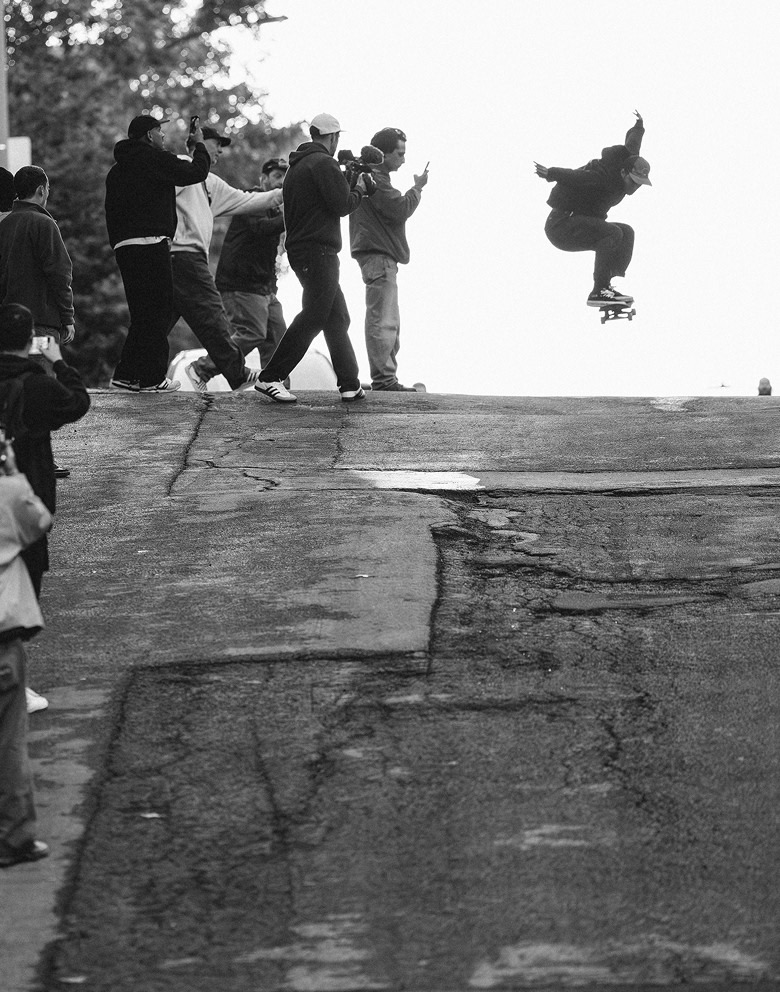 A black and white picture of a skater catching air on a downhill street as people watch.
