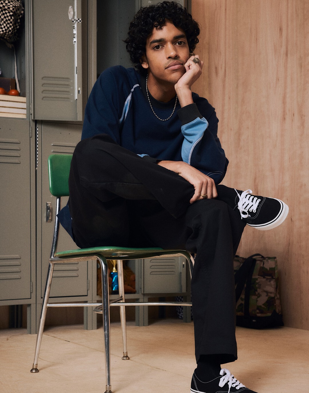 A young man poses against a wooden background next to school lockers wearing Vans shoes, clothes, and a backpack.