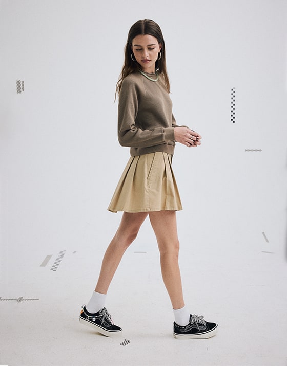 A young woman wearing an incense brown Authentic Chino Mini Skirt and a bungee cord brown Cabrillo Long Sleeve Crew Sweatshirt in a photo studio.