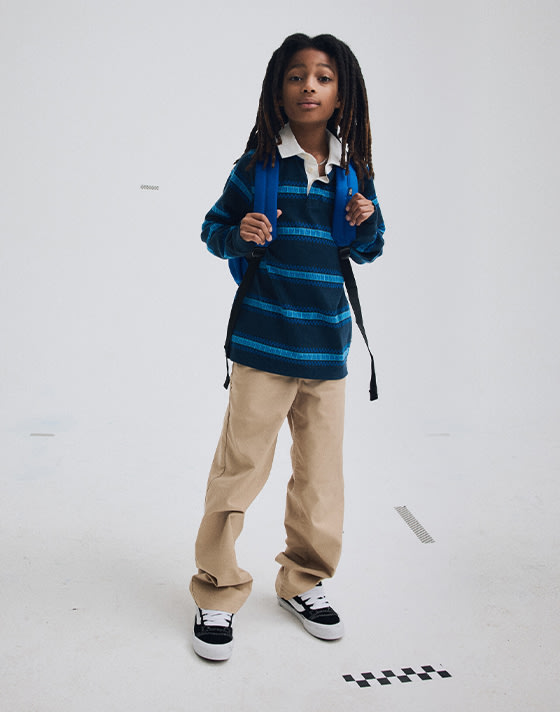 A young boy poses in a photo studio wearing black and white Knu Skool, Kids Authentic Chino Loose Pants, and a Kids V Rugby Long Sleeve Shirt.