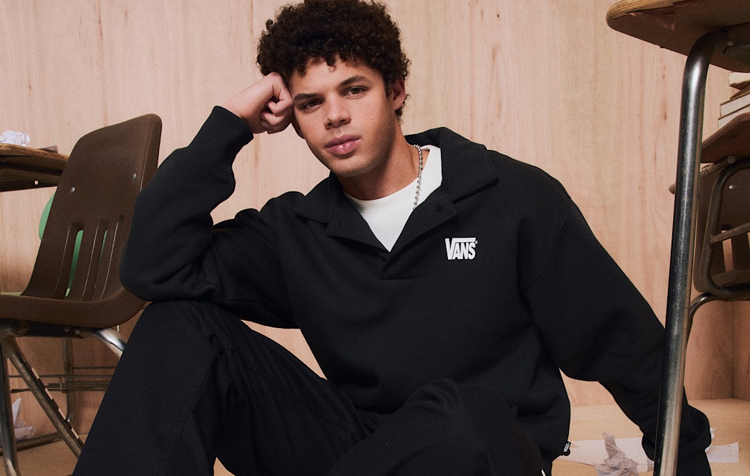 A young man sits on a classroom floor and looks into the camera wearing Vans shoes and clothes.