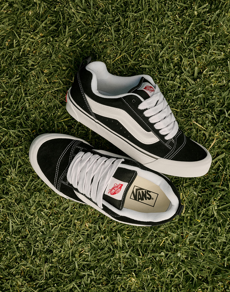 An overhead shot of a pair of black and white Knu Skool lie on the grass at golden hour.