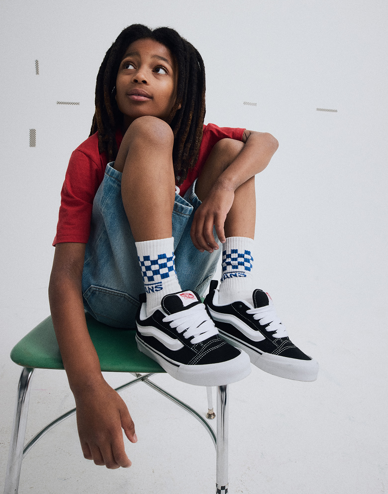 Medium shot of a kid sitting on a chair while wearing a pair of black / white Knu Skool shoes, blue checkerboard socks, jean shorts and a red shirt.