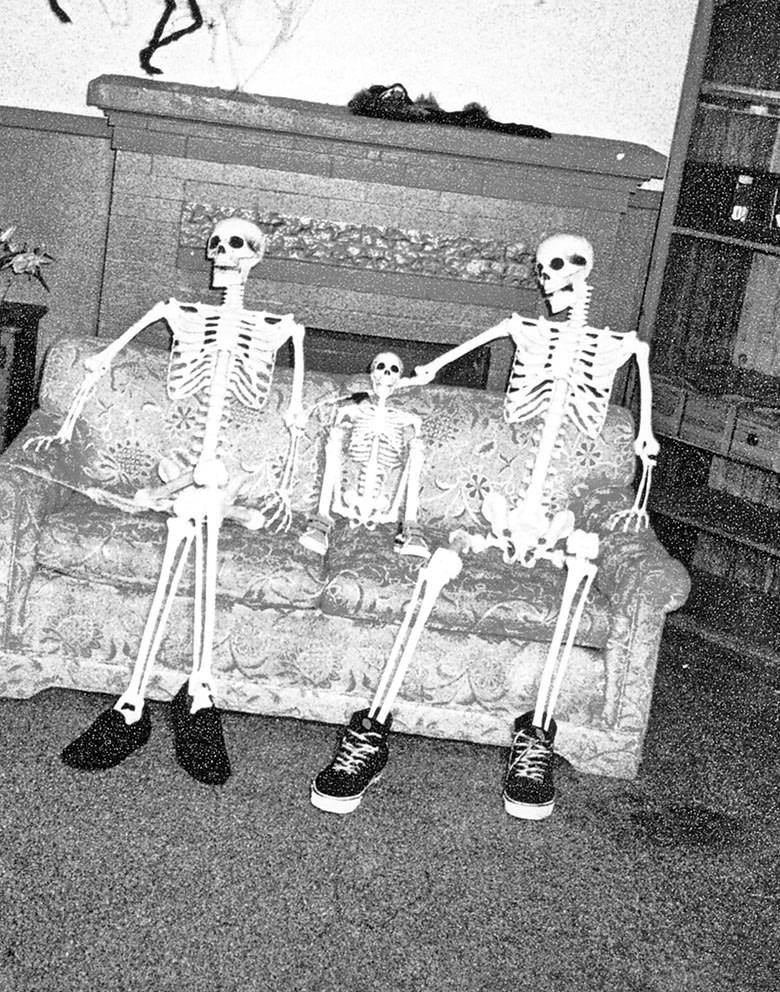 Black and white photo. Wide shot of a couch in an old house with three skeletons sitting on it. Smaller one in the middle, two larger ones beside it.
