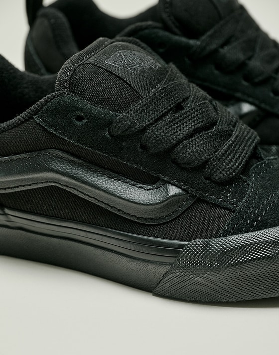 Close-up on a pair of all black Knu Skool on the floor of a photography studio.