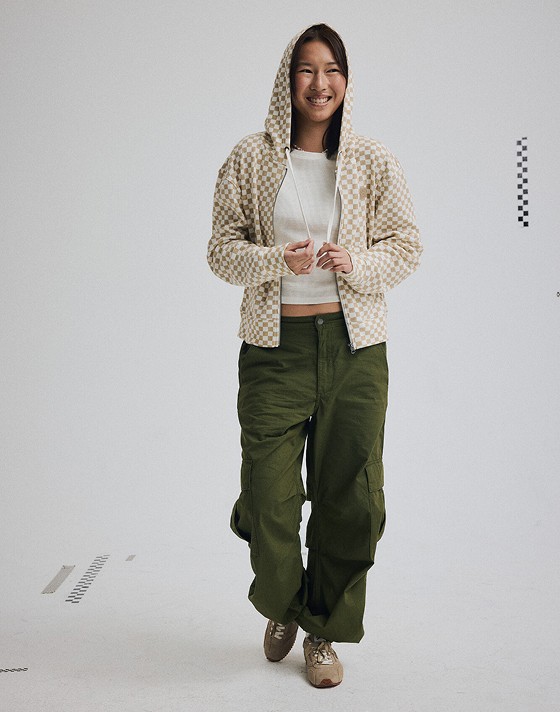 A young woman poses in a photo studio wearing green Para Baggy Cargo Pants with a brown and white Checkerboard Pierce Shrunken Full Zip.