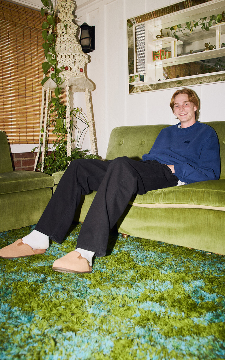A young man sits on a living room couch wearing a pair of incense brown Harbor Mule VR3 and new Vans styles.