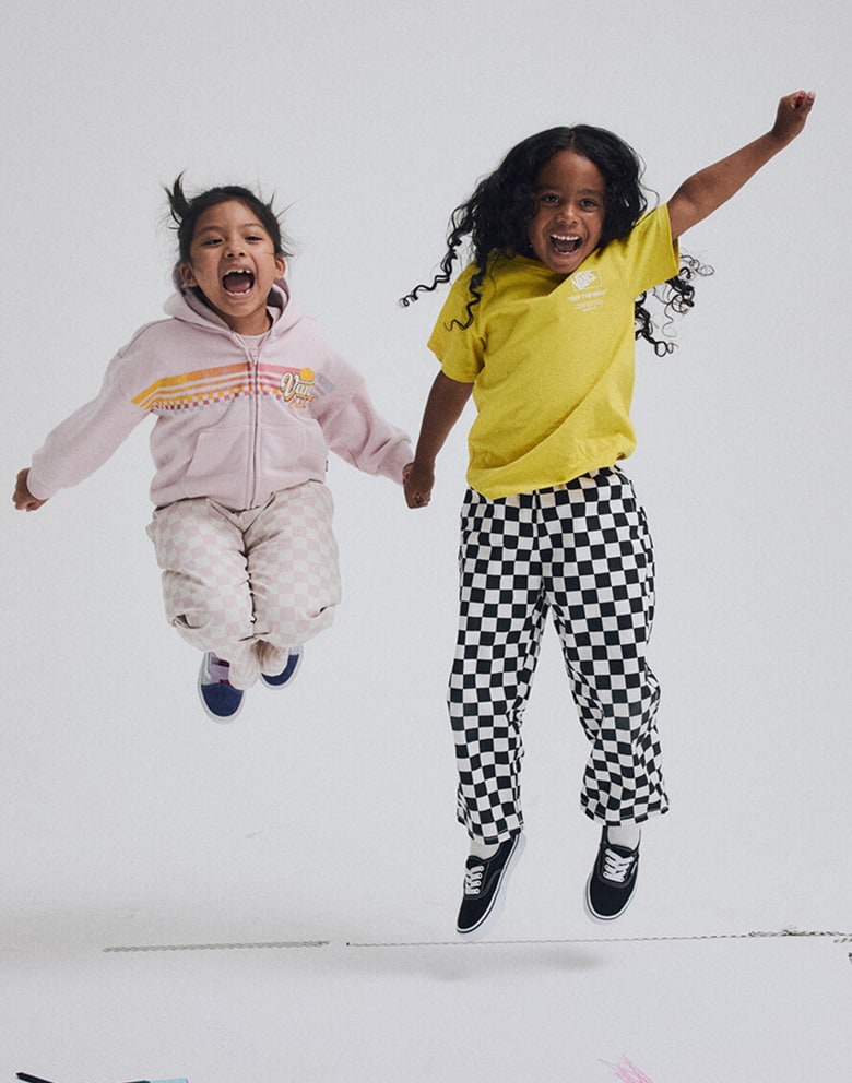 A young boy and girl jump together and smile at the camera wearing new Vans Little Kids styles.