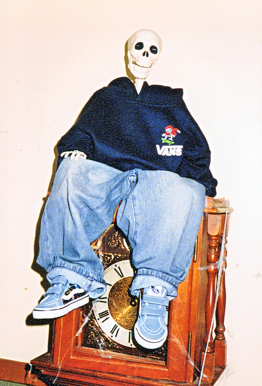 Skeleton figure wearing a Vans graphic hoodie, baggy jeans, and blue Vans easy-on, easy-off sneakers seated on a vintage grandfather clock decorated with cobwebs.