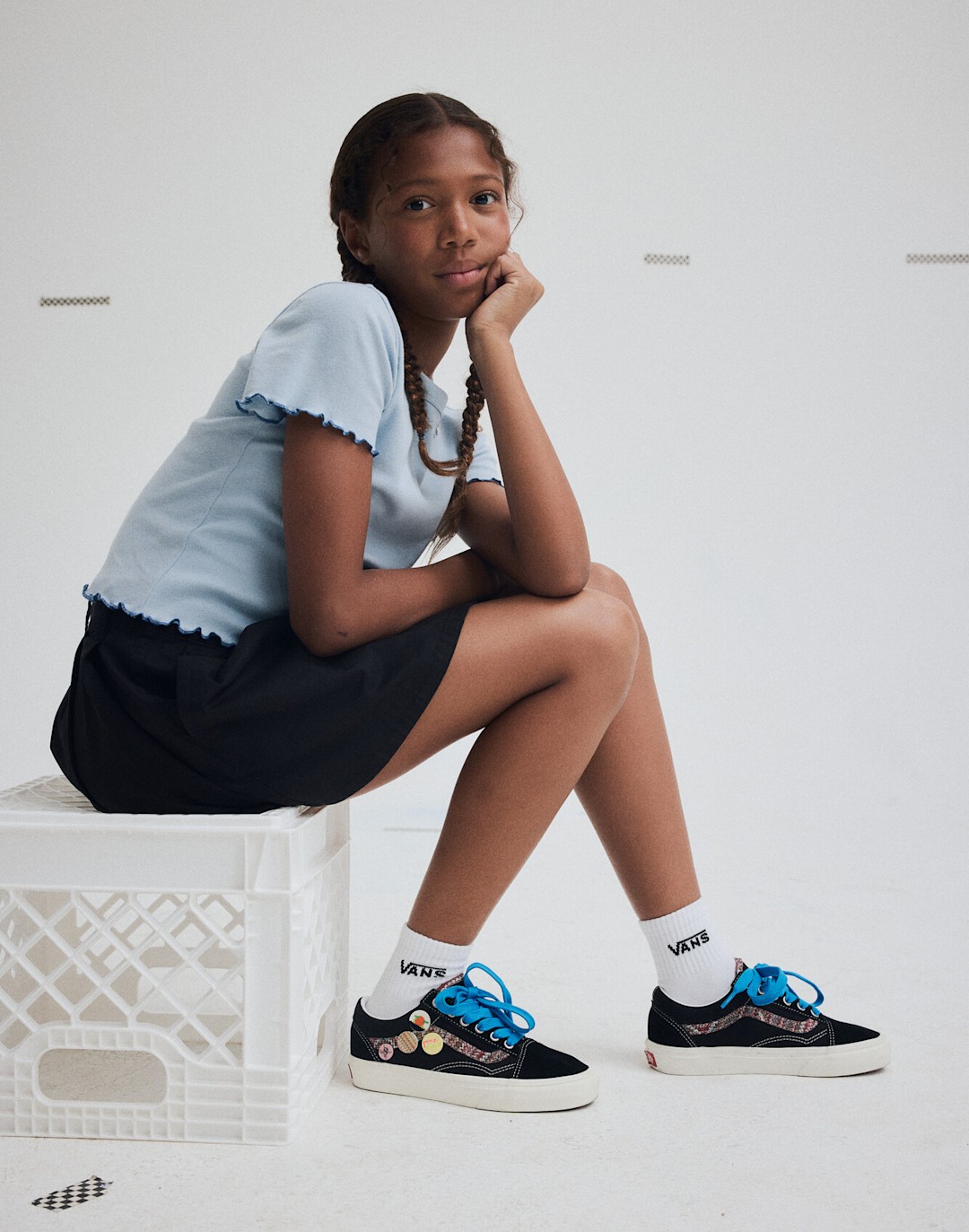 A young girl sits on a milk crate, looking at the camera, wearing Vans clothing and shoes.