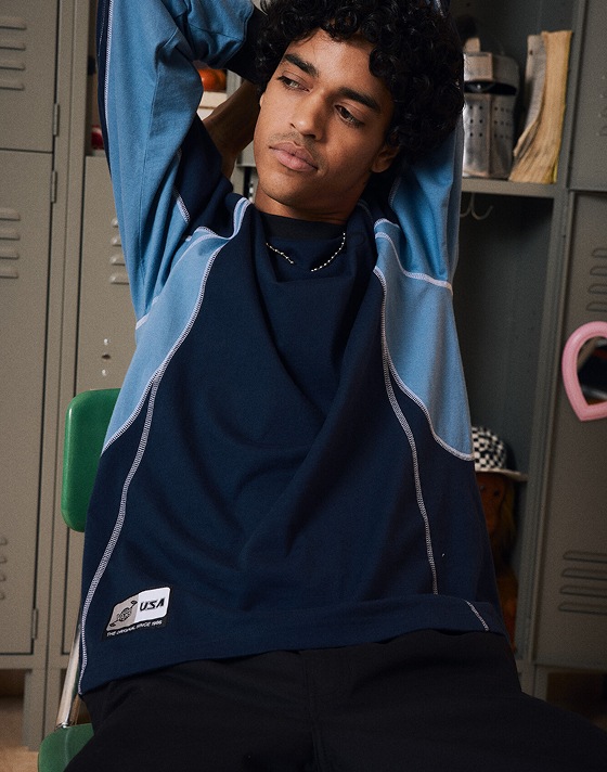 Waist up close up of a man sitting in a chair with lockers behind him. He’s wearing the Retro Sport Long Sleeve T-Shirt in Dress Blues.