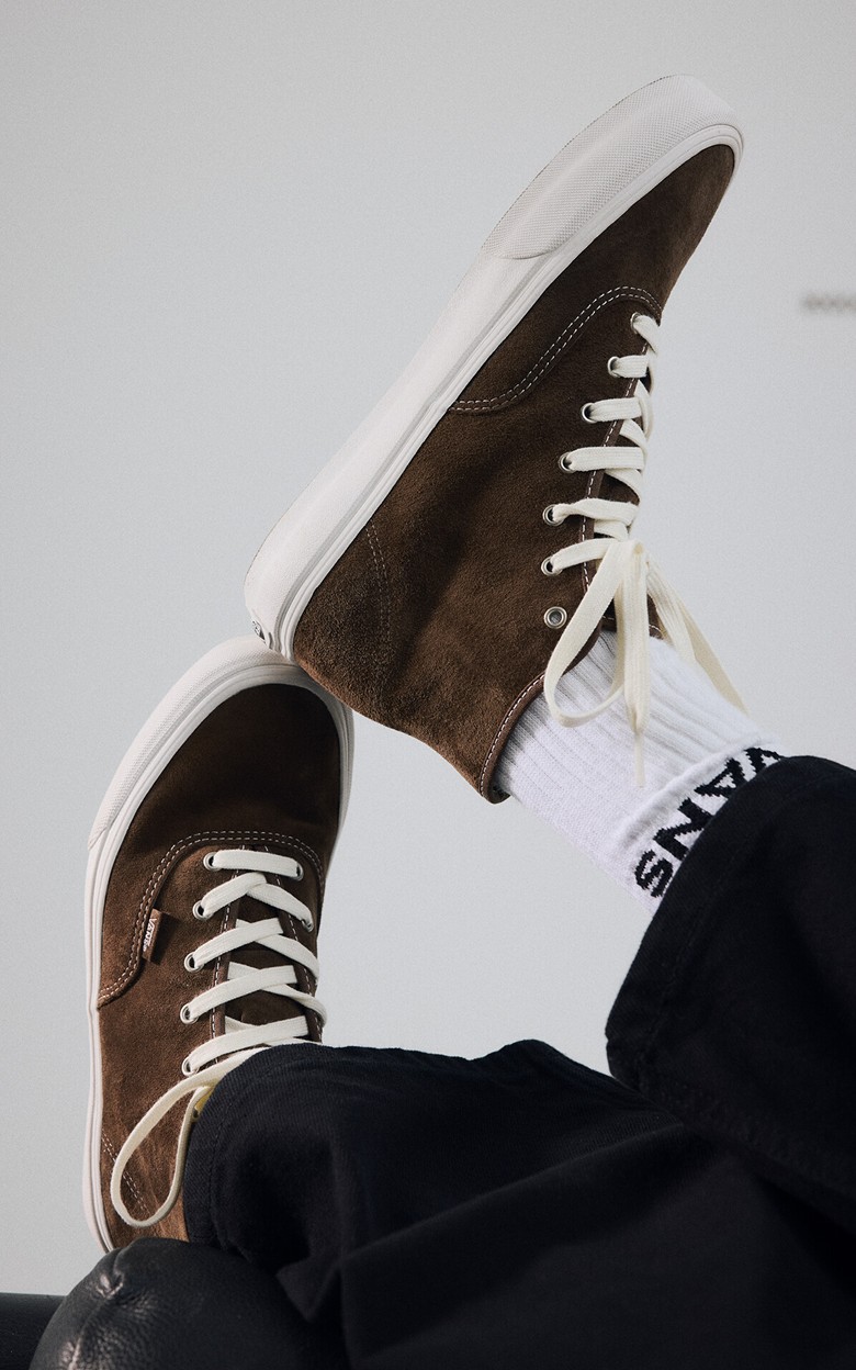 Close-up on someone kicking back and stacking their brown suede Authentic Hi 2.0 shoes on top of each other.