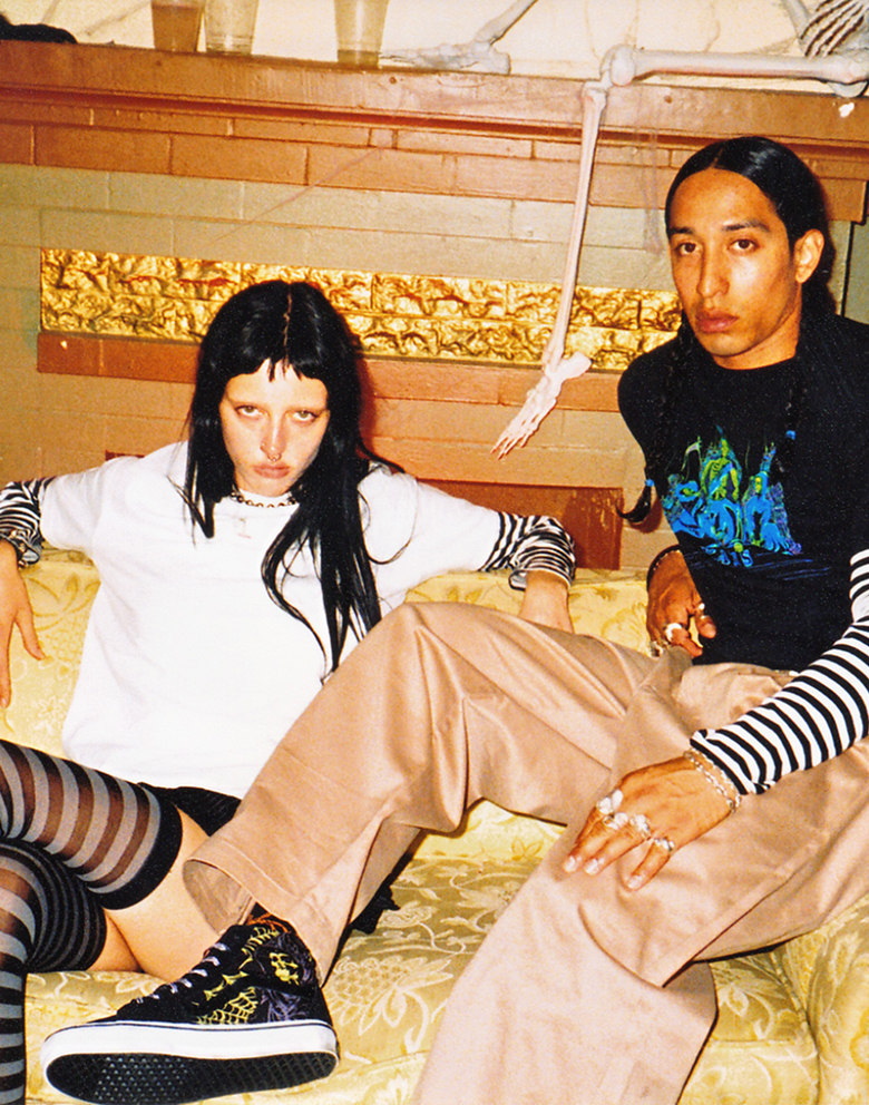 Wide shot of two people sitting on a couch in an old spooky living room with a skeleton behind them on the mantle. Both dressed in halloween styles.