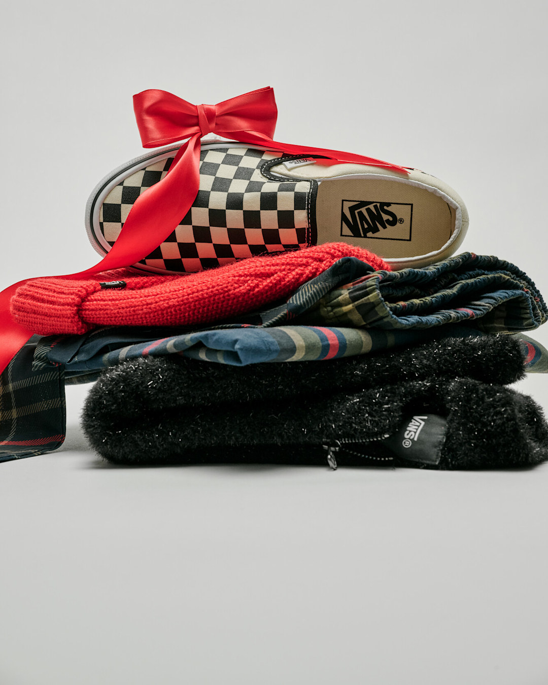 Checkerboard Vans Slip-On shoe wrapped with a red ribbon and bow, displayed on top of folded holiday apparel and accessories.