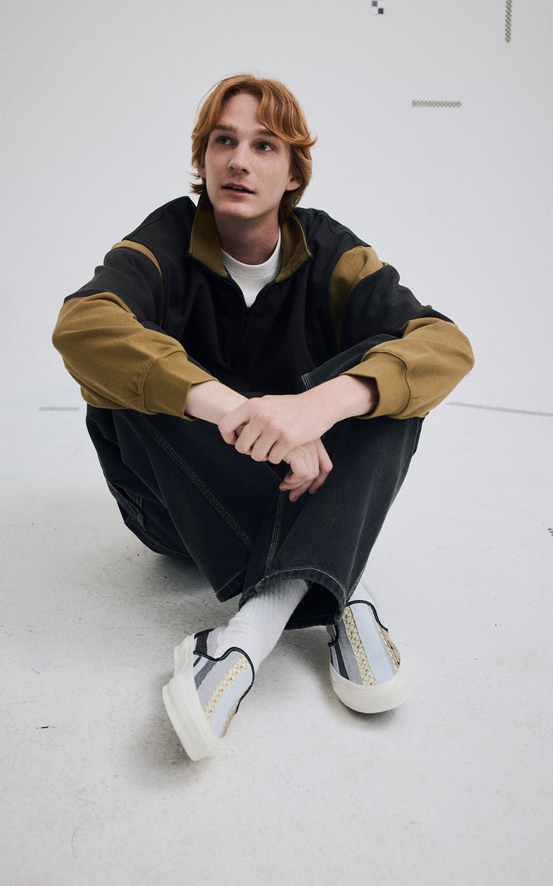 Medium shot of a man sitting down in a grey photography studio wearing the Premium Classic Slip-On Shoe in Blue / Black with jeans and a rugby shirt.