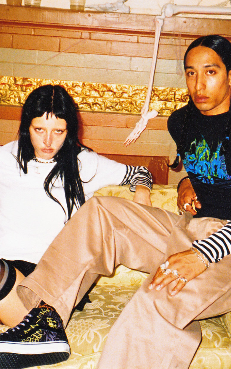 Wide shot of two people sitting on a couch in an old spooky living room with a skeleton behind them on the mantle. Both dressed in halloween styles.