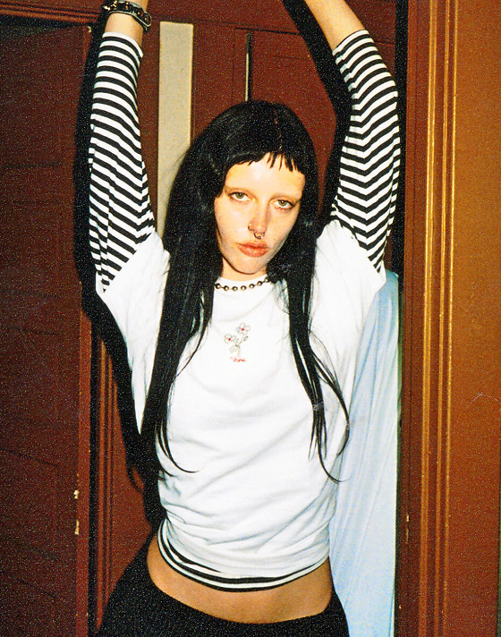 Wide shot of a woman in an old house wearing a black and white long sleeve shirt with another shirt with flowers over the top.