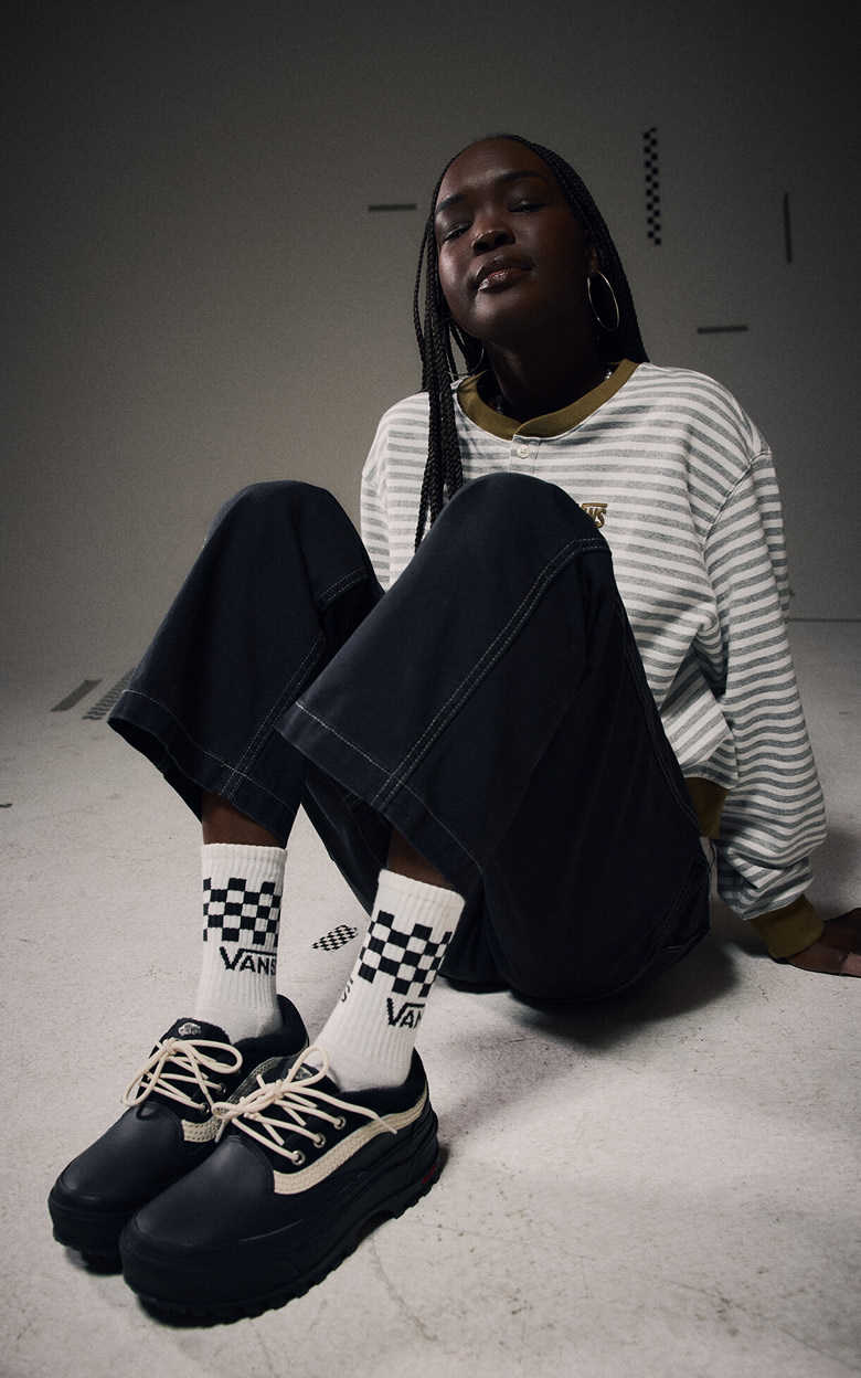 Medium shot of a woman in a dark gray photography studio wearing a long-sleeve shirt, jeans, checkered socks, and the Standard Low in black.