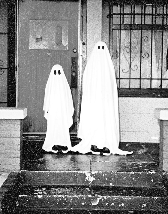 Black and white medium shot of two kids with sheets over themselves with eye holes cut out to look like ghosts in front of a creepy house.