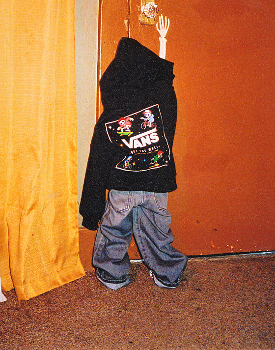 Medium shot of a little skeleton facing the door, reaching for the handle while wearing a Little Kids Glow Bones Pullover Hoodie and dark jeans.