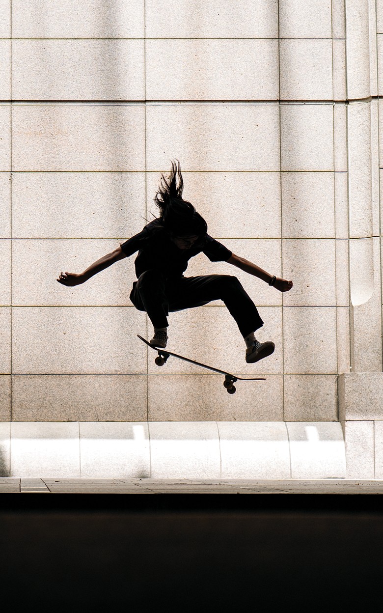 A stylized photo of a skater covered in shadow performing an ollie outside of an office building on the sidewalk.