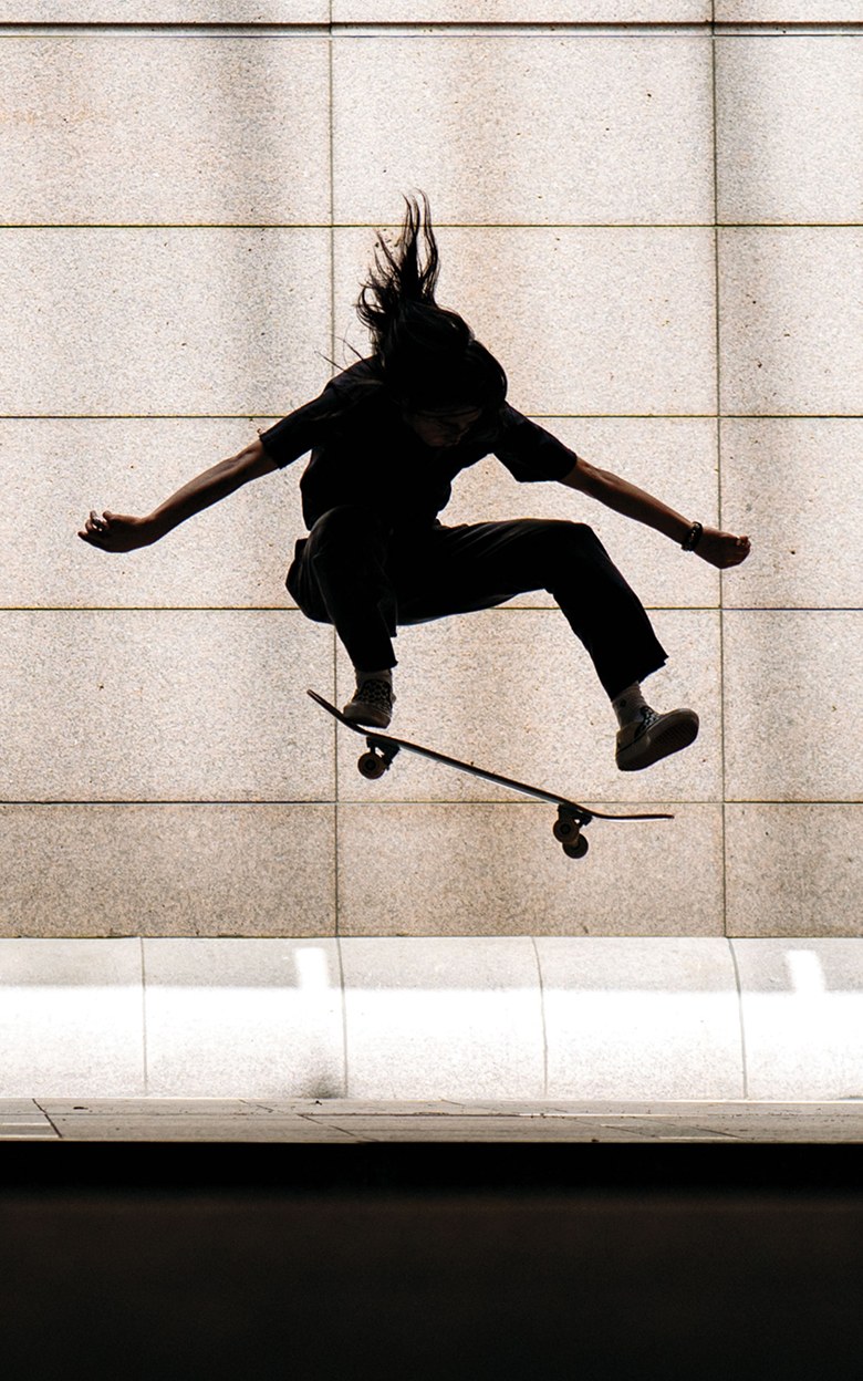 A stylized photo of a skater covered in shadow performing an ollie outside of an office building on the sidewalk.