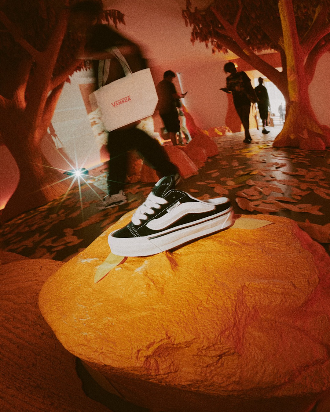 Classic black-and-white Vans sneaker displayed on a raised platform within an immersive exhibit featuring tree-inspired structures, leaf-covered flooring, and warm ambient lighting. Attendees walk through the space in the background.