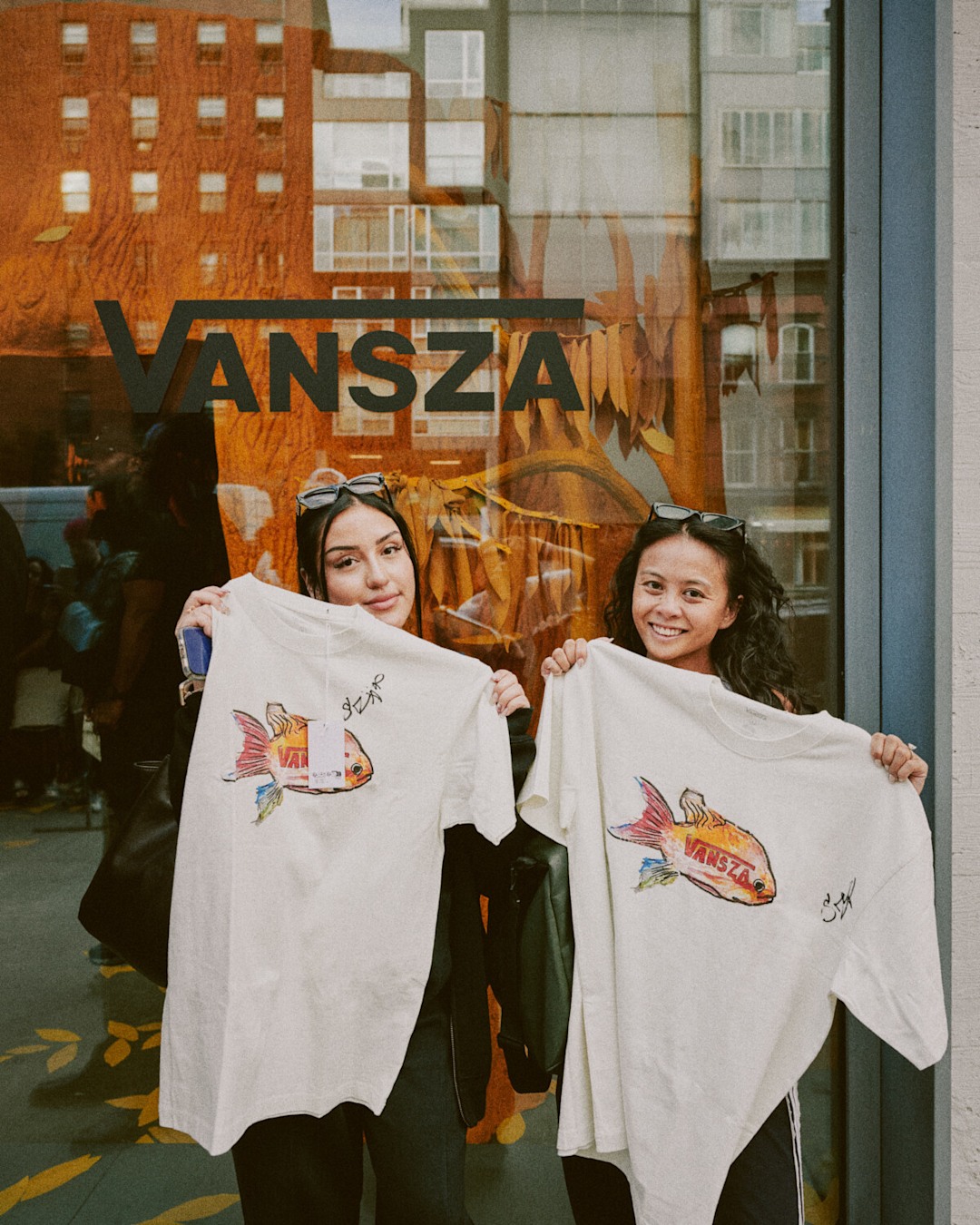 Two people stand outside a storefront holding white graphic T-shirts. A large storefront window with the text “VANSZA” is visible in the background, along with reflections of nearby buildings.