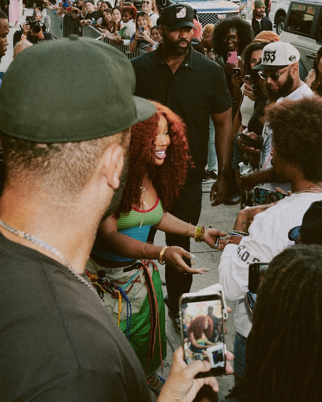 Person interacting with a crowd at an outdoor event while wearing a colorful striped top and green shorts. Attendees surround the scene, taking photos and recording on mobile phones.
