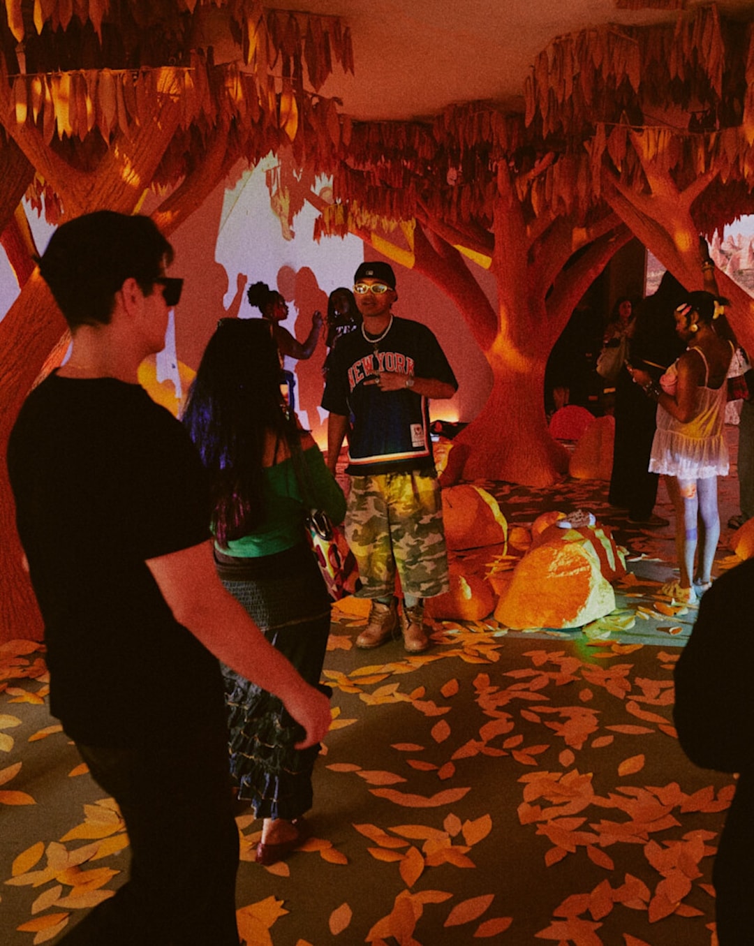 Indoor event space featuring large tree-inspired sculptures, leaf-shaped decorations across the floor and ceiling, and warm orange lighting. Multiple attendees are gathered throughout the exhibit exploring the themed environment.