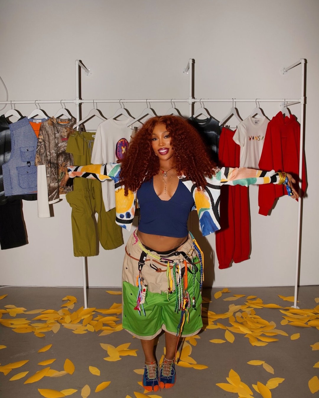 Person standing with arms outstretched in front of a clothing rack displaying apparel and footwear. Wearing a blue crop top, multicolored jacket, and green shorts with decorative details. Yellow leaf shapes are scattered across the floor.