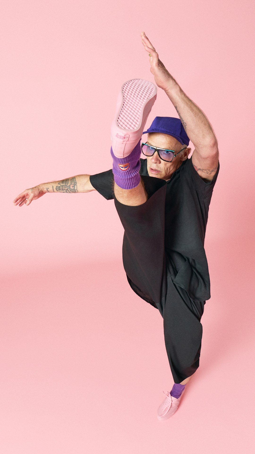 A person dressed in black poses against a soft pink background, raising one leg high to showcase the waffle outsole of a pink Vans Authentic shoe. Purple socks and a matching purple cap contrast with the pink footwear and backdrop, creating a bold, studio-style composition.